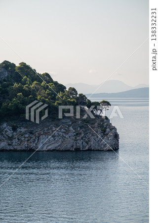 Rocky forested cliff above calm sea with distant mountains 138332231