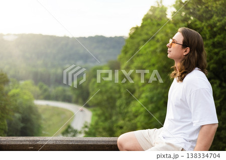 Profile of a relaxed young man with long hair and sunglasses sitting on a stone wall, enjoying a scenic view of a winding road and lush green forest at sunset. Profile of a relaxed young man with long hair and sunglasses sitting on a stone wall, enjoying a scenic view of a winding road and lush green forest at sunset. 138334704