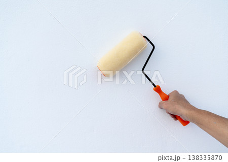 A person's hand holding a paint roller against a clean white textured wall. Home renovation and interior improvement concept with a yellow roller and orange handle on a bright background. 138335870