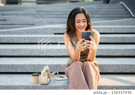 Pretty asian woman smiling at phone sitting with bag and coffee on concrete stairs in park or plaza. Pretty asian woman smiling at phone sitting with bag and coffee on concrete stairs in park or plaza. 138337436