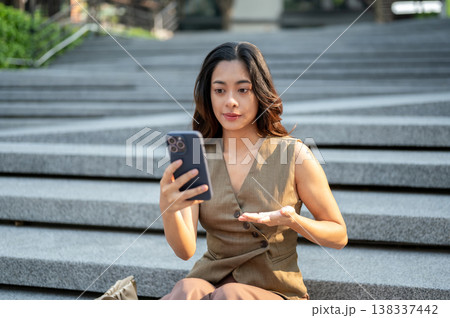 Pretty asian woman hand explaining talking on video phone call as sitting on concrete stairs in park Pretty asian woman hand explaining talking on video phone call as sitting on concrete stairs in park 138337442