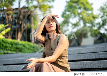 Pretty asian woman using a hand covering eyes from sunlight while sitting on concrete stairs in park 138337449