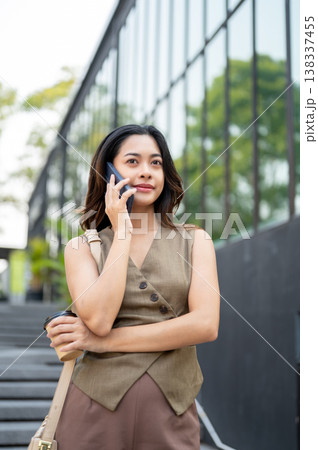 Pretty asian woman talking on phone folding arm holding coffee while standing or walking by building Pretty asian woman talking on phone folding arm holding coffee while standing or walking by building 138337455