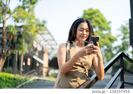 Pretty asian woman typing or looking at smartphone while standing at stairs's railing in park plaza. Pretty asian woman typing or looking at smartphone while standing at stairs's railing in park plaza. 138337458