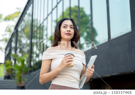 Pretty asian woman carrying a laptop and coffee cup while standing or walking on stairs by building. 138337468