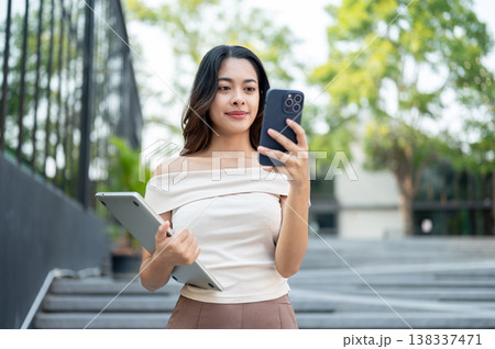 Pretty asian woman looking at phone holding a laptop while standing on concrete stairs by building. 138337471