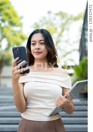 Pretty asian woman looking at phone holding a laptop while walking down concrete stairs by building. 138337472