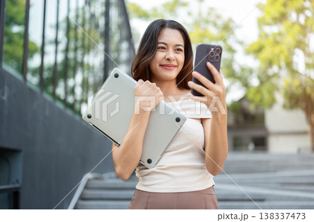 Happy asian woman looking at phone holding a laptop while walking down concrete stairs by building. 138337473