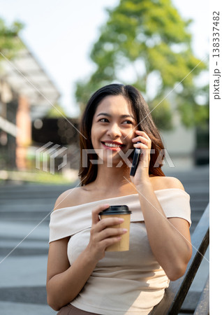 Pretty asian woman talking on phone call holding coffee while standing leaning on a railing in park. Pretty asian woman talking on phone call holding coffee while standing leaning on a railing in park. 138337482