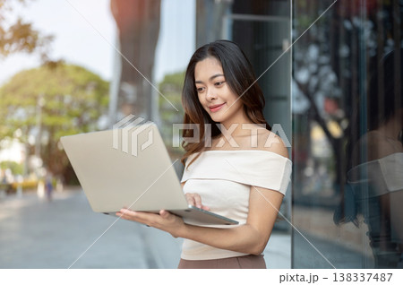 Pretty asian woman student freelancer looking at laptop while standing or walking outside a building Pretty asian woman student freelancer looking at laptop while standing or walking outside a building 138337487