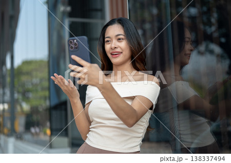 Pretty asian woman holding phone talking on video call standing at glass wall outside mall building. Pretty asian woman holding phone talking on video call standing at glass wall outside mall building. 138337494