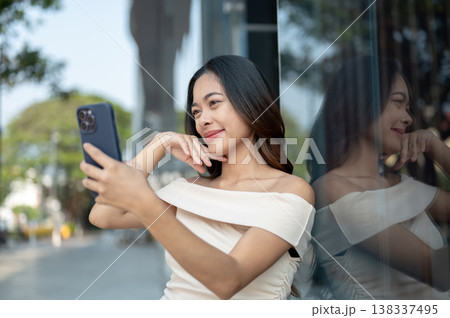 Pretty asian woman holding phone taking selfie picture standing at glass wall outside mall building. 138337495