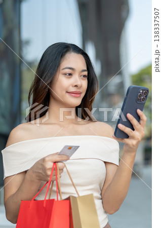 Asian woman customer looking at phone holding credit card with shopping bags standing outside a mall Asian woman customer looking at phone holding credit card with shopping bags standing outside a mall 138337507