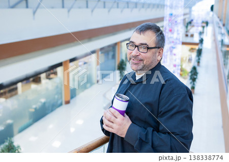 Man holding takeaway coffee in modern mall interior. Man in dark clothing sitting near balcony holding takeaway cup inside bright shopping mall with glass ceiling and greenery Man holding takeaway coffee in modern mall interior. Man in dark clothing sitting near balcony holding takeaway cup inside bright shopping mall with glass ceiling and greenery 138338774