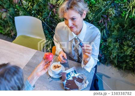 Two mature women sitting at wooden table in modern cafe holding cups of coffee and dessert engaged in friendly conversation. Two mature women sitting at wooden table in modern cafe holding cups of coffee and dessert engaged in friendly conversation. 138338782