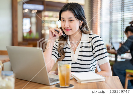 Pretty asian woman student holding pen aside face looking at laptop with book an drink on cafe table Pretty asian woman student holding pen aside face looking at laptop with book an drink on cafe table 138339747