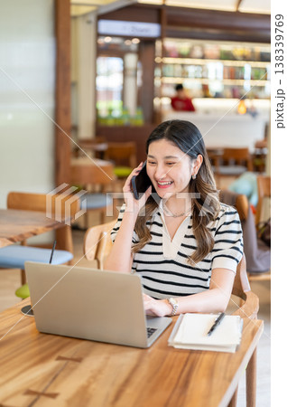 Pretty asian woman talking on phone looking at laptop aside notebook while sitting at table in cafe. 138339769