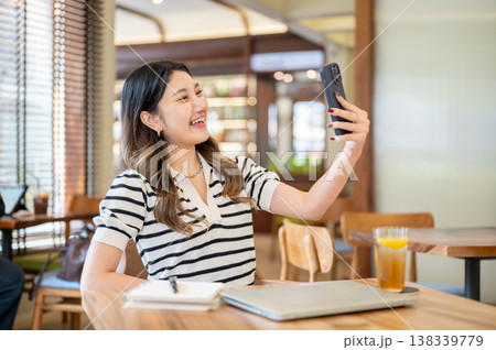 Pretty asian woman holding phone talking on video call aside laptop and notebook at table in cafe. Pretty asian woman holding phone talking on video call aside laptop and notebook at table in cafe. 138339779