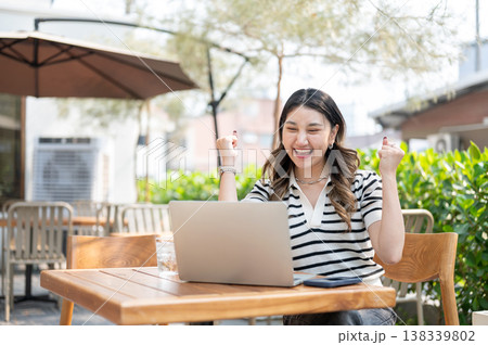 Excited asian woman looking at laptop and making a fist while sitting at wooden table in garden cafe 138339802