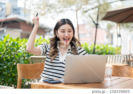 Excited asian woman looking at laptop as raising a fist while sitting at wooden table in garden cafe 138339803
