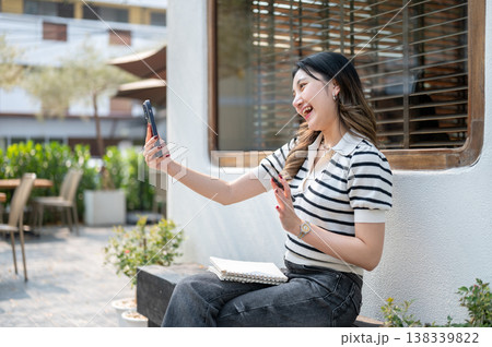 Pretty asian woman holding phone talking on video call while sitting on wooden bench in garden cafe. Pretty asian woman holding phone talking on video call while sitting on wooden bench in garden cafe. 138339822