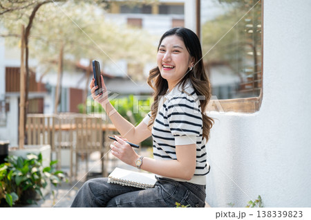 Pretty asian woman holding pen and phone over notebook while sitting on wooden bench in garden cafe. Pretty asian woman holding pen and phone over notebook while sitting on wooden bench in garden cafe. 138339823