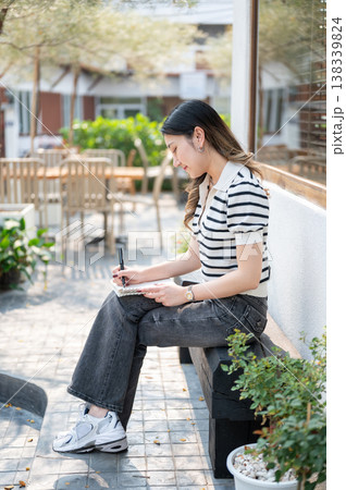 Asian woman holding pen writing in notebook and sitting cross legged on wooden bench in garden cafe. 138339824