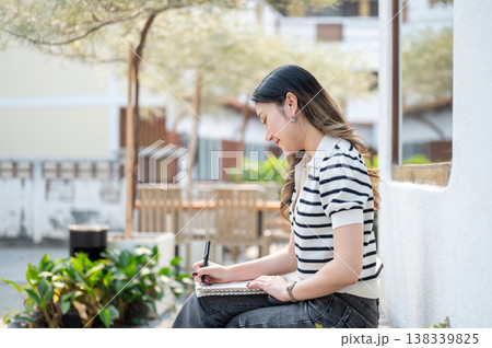 Asian woman holding a pen writing in notebook as sitting cross legged on wooden bench in garden cafe Asian woman holding a pen writing in notebook as sitting cross legged on wooden bench in garden cafe 138339825