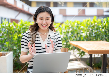 Pretty asian woman talking on video call with a laptop while sitting on wooden chair in garden cafe. Pretty asian woman talking on video call with a laptop while sitting on wooden chair in garden cafe. 138339829