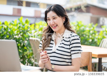 Happy asian woman clasping hands talking and looking at laptop while sitting at table in garden cafe Happy asian woman clasping hands talking and looking at laptop while sitting at table in garden cafe 138339835