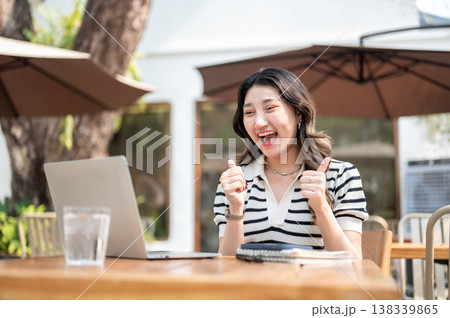 Happy asian woman giving thumbs up as looking at laptop while sitting at wooden table in garden cafe 138339865