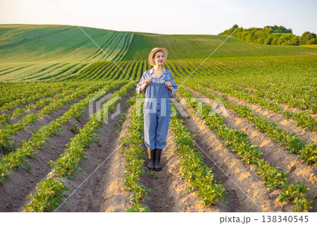 A young woman wearing overalls and a straw hat stands in a field of young crops with rolling green hills in the background A young woman wearing overalls and a straw hat stands in a field of young crops with rolling green hills in the background 138340545