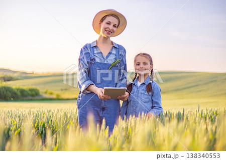 A woman in overalls and a straw hat holds a tablet while standing with her daughter in a sunlit wheat field, representing modern agriculture and family 138340553