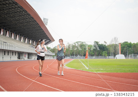 Athletic asian woman friends talking while running jogging together on racetrack in sports stadium. Athletic asian woman friends talking while running jogging together on racetrack in sports stadium. 138343215