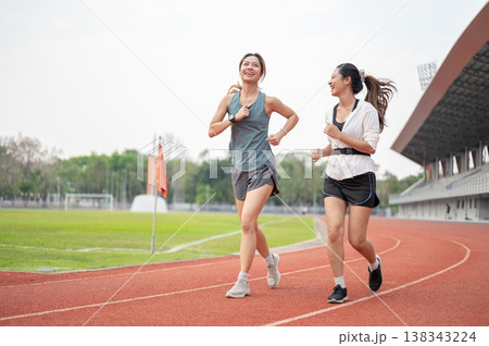 Athletic asian woman and her friend talking together while running on racetrack in sports stadium. Athletic asian woman and her friend talking together while running on racetrack in sports stadium. 138343224