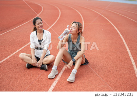 Asian woman sitting on racetrack aside a friend drinking water while resting after running exercise 138343233