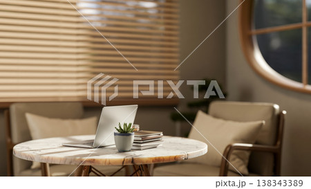 Laptop and books on round marble table across wooden blinds and armchair in the living room or cafe. Laptop and books on round marble table across wooden blinds and armchair in the living room or cafe. 138343389