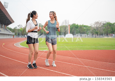 Pretty asian woman friends talking while running or jogging together on racetrack in sports stadium. Pretty asian woman friends talking while running or jogging together on racetrack in sports stadium. 138343565