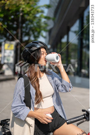 Happy asian woman carrying eco bag drinking water from tumbler bottle while riding a bicycle in park 138343733