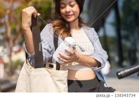 Close up asian woman putting thermos bottle in eco bag while riding a bicycle in park under sunlight Close up asian woman putting thermos bottle in eco bag while riding a bicycle in park under sunlight 138343734