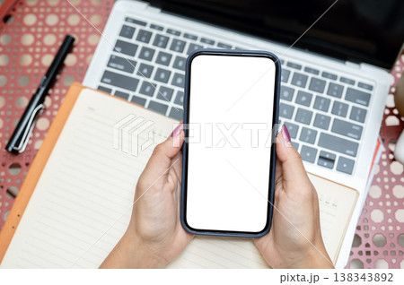 Top view of a woman's hands holding white screen smartphone over book and laptop at table in cafe. Top view of a woman's hands holding white screen smartphone over book and laptop at table in cafe. 138343892