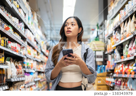 Asian woman holding phone looking at product shelf while shopping or buying grocery in supermarket. Asian woman holding phone looking at product shelf while shopping or buying grocery in supermarket. 138343979