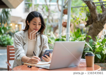 Asian businesswoman or student looking at phone writing aside laptop sitting at table in garden cafe 138343981