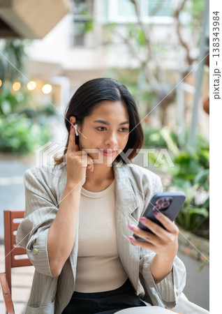 Pretty asian woman holding earphone listening to music making a call sitting at table in garden cafe Pretty asian woman holding earphone listening to music making a call sitting at table in garden cafe 138343984