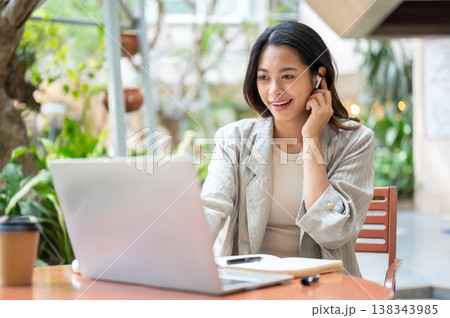 Pretty asian woman wearing earphone talking on call across laptop as sitting at table in garden cafe Pretty asian woman wearing earphone talking on call across laptop as sitting at table in garden cafe 138343985