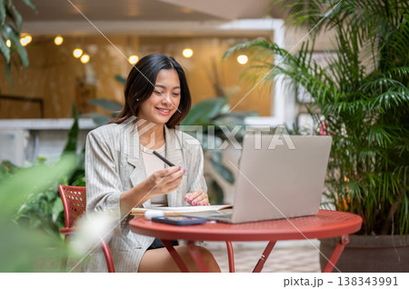 Pretty asian woman student or freelancer holding pen writing aside a laptop at table in garden cafe. Pretty asian woman student or freelancer holding pen writing aside a laptop at table in garden cafe. 138343991