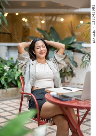 Pretty asian woman student sitting with hands behind head looking at laptop on table in garden cafe. Pretty asian woman student sitting with hands behind head looking at laptop on table in garden cafe. 138343993