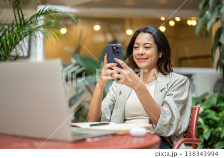 Pretty asian woman student looking at phone over book and laptop as sitting at table in garden cafe. Pretty asian woman student looking at phone over book and laptop as sitting at table in garden cafe. 138343994