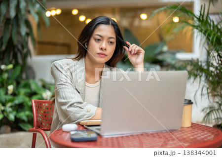 Pretty asian businesswoman holding pen touching head looking at laptop sits at table in garden cafe. Pretty asian businesswoman holding pen touching head looking at laptop sits at table in garden cafe. 138344001