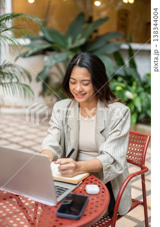 Happy asian businesswoman or student holding pen writing aside laptop sits at table in garden cafe. Happy asian businesswoman or student holding pen writing aside laptop sits at table in garden cafe. 138344004